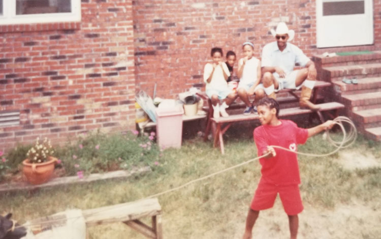 A home photo of a young Candace Dantes roping the dummy and her family sitting watching her.